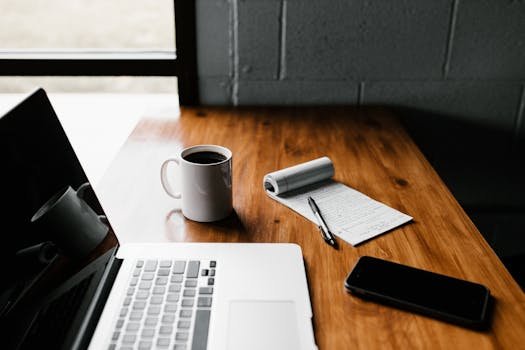 A tidy workspace featuring a laptop, coffee cup, phone, and notepad on a wooden desk.